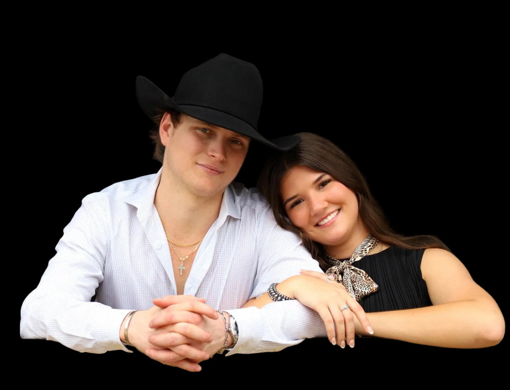 A smiling young couple posing together outdoors under a rustic covered structure. The man wears a black cowboy hat, a white button-down shirt, and gold jewelry.