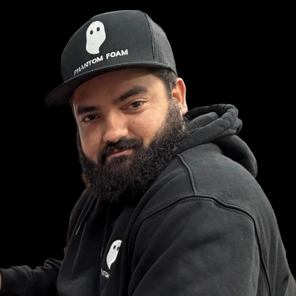 A man with a dark beard wearing a black Phantom Foam trucker hat and a black hoodie, photographed in an office or retail environment.