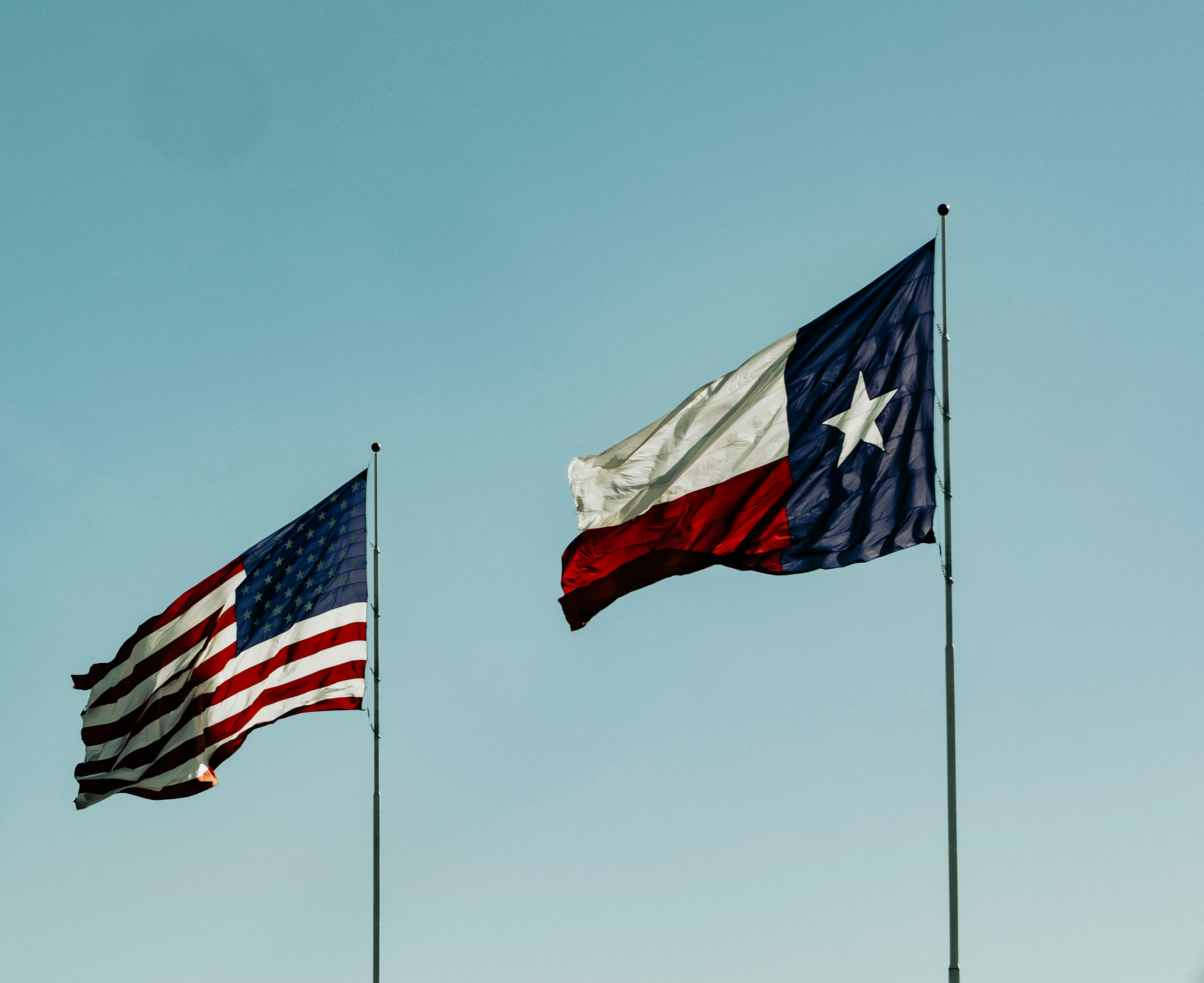 American flag and Texas flag waving side by side against a clear blue sky.
